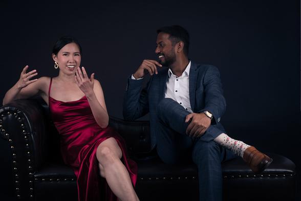 Zwei Personen: Links sitzt eine asiatische Frau im rot-glänzenden langen Kleid und gestikuliert mit den Händen. Sie blickt in die Kamera. Rechts sitzt ein Mann mit dunkler Haut im blauen Anzug, greift sich ans Kinn. Er hört ihr lachend zu. Gut sichtbar sind seine weißen Socken mit Melonenscheibenaufdruck.