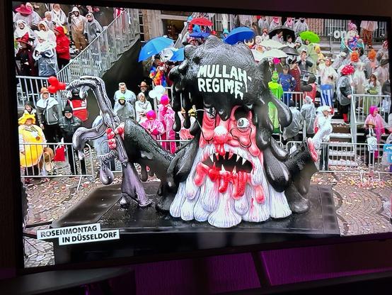 Ein großer, grotesker Wagen, der ein Monster mit übertriebenen Gesichtszügen, einschließlich scharfer Zähne und einer wilden Frisur, darstellt, ist in einer Parade-Umgebung zu sehen. Auf dem Schwimmer wird der Text "MULLAH-REGIME" prominent angezeigt. Im Hintergrund eine Menschenmenge.

A large, grotesque float depicting a monster with exaggerated features, including sharp teeth and a wild hairstyle, is featured in a parade setting. The float has the text "MULLAH-REGIME" prominently displayed. In the background, a crowd.