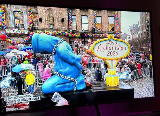 Eine farbenfrohe Paradeszene mit einem großen Wagen. Der Wagen zeigt eine blaue Figur in Ketten mit einem Schild mit der Aufschrift "Miss Afghanistan 2026". Im Hintergrund versammelt sich eine Menschenmenge unter Regenschirmen, und die Kulisse erscheint festlich mit Luftballons und Dekorationen.

A colorful parade scene featuring a large float. The float displays a blue figure in chains with a sign reading "Miss Afghanistan 2026." In the background, a crowd is gathered under umbrellas, and the setting appears festive with balloons and decorations.