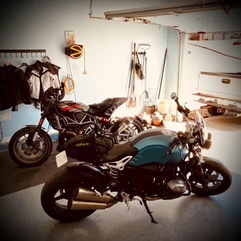 Two motorcycles parked in the garage.  In the foreground is a late model BMW RnineT facing right towards the garage door that is open and letting in all the sunshine. Behind it, facing the opposite direction, is a late model Indian FTR 1200.  
On the wall behind the bikes hangs a rack of motorcycle jackets and gear