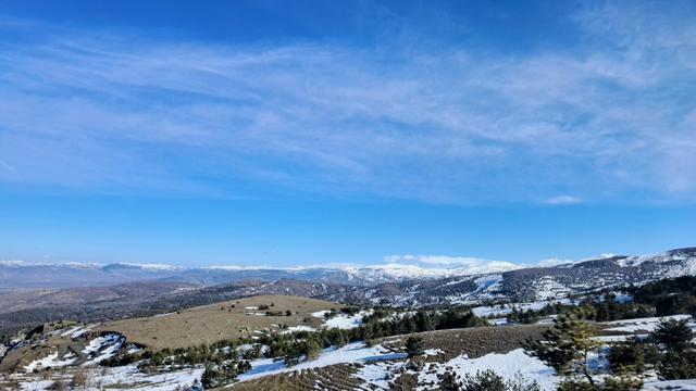 A wide landscape view of rolling hills partially covered in snow and scattered pine trees. In the distance, a snow-capped mountain range stretches across the horizon, with what appear to be faint wind turbines visible on the far left ridge. A bright blue sky with wispy white clouds fills the upper half of the scene. powershell new-guid Guid 77948837-bb70-4c12-8611-cb3f4d252f3f