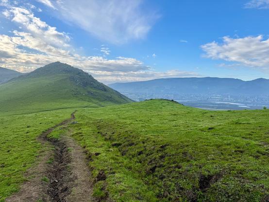 A muddy trail cuts through green hills and valleys. There is another large hill ahead, and blue mountains in the distance under a partly blue sky with white and grey clouds. It's a somewhat minimalist landscape and image. This is in the Laguna Lake Open Space at the base of Cerro San Luis mountain in San Luis Obispo, California.