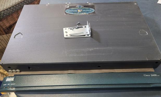 A large dark grey metal box with three LEDs on the front and a dark green faceplate lying in front of it. The faceplate has the Cisco Systems logo, labels for the LEDs, and Cisco 2600 printed on it. A pair of rack mounting ears and four screws are lying no top of it.