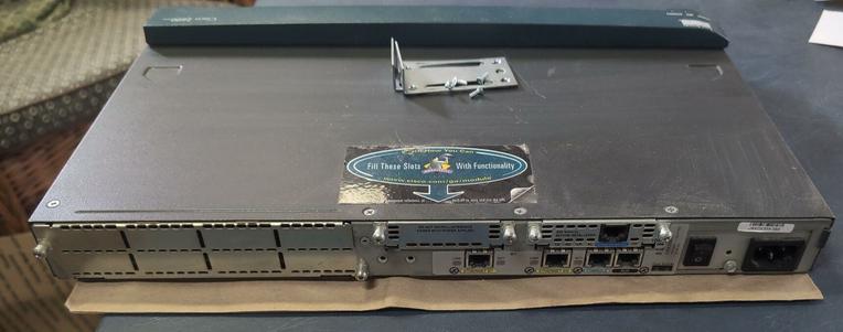 A shot of the router from the back. A large blanking panel covers the left half. A small blanking panel is in the middle. A large sticker on the top with an arrow to the blanking panel says "Fill these slots with functionality." Below and to the right are modular jacks, a power switch, and IEC inlet.