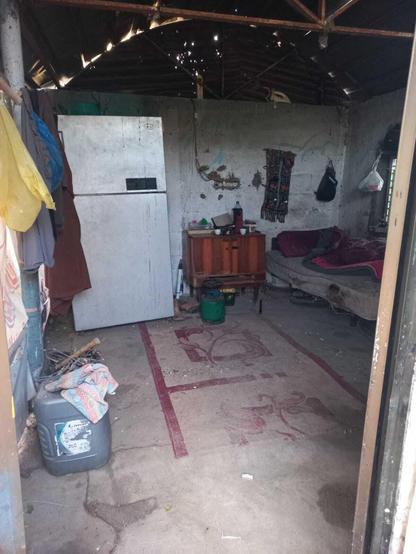 Interior of a dilapidated room with a refrigerator, rug, bed, and small wooden cabinet, visible through a doorway.