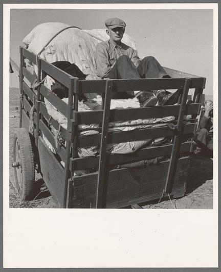 A black and white photograph shows an individual sitting in a wagon filled with various items, including blankets or tarps. The person is wearing casual work attire: a cap, striped shirt, dark trousers, and boots. They appear to be resting on the contents of the wagon during what could be considered a laborious journey across dry terrain under clear skies. This scene reflects rural life's hardships, likely from an early 20th-century American context given the style of clothing and type of cart used for transportation.