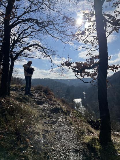 Eine Person steht auf einem Hügel, umgeben von Bäumen, und schaut auf einen gewundenen Fluss darunter. Der Himmel ist teilweise bewölkt, wobei Sonnenlicht durchbricht und eine ruhige Außenszene entsteht.