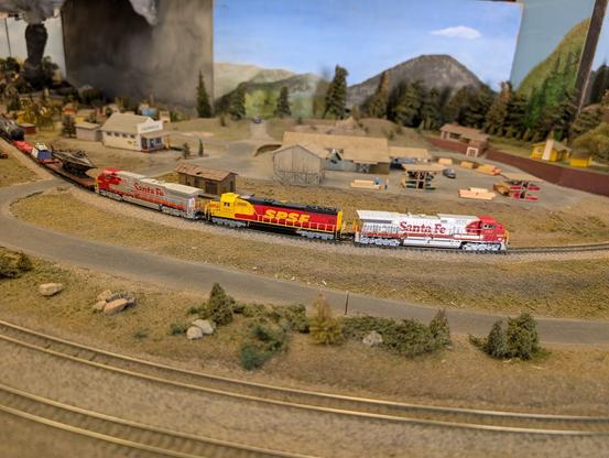 3 locomotives pull a train around the corner on an N-scale model railroad layout. The lead and trailing engines are Santa Fe Warbonnets, with the middle engine being a SPSF Kodachrome