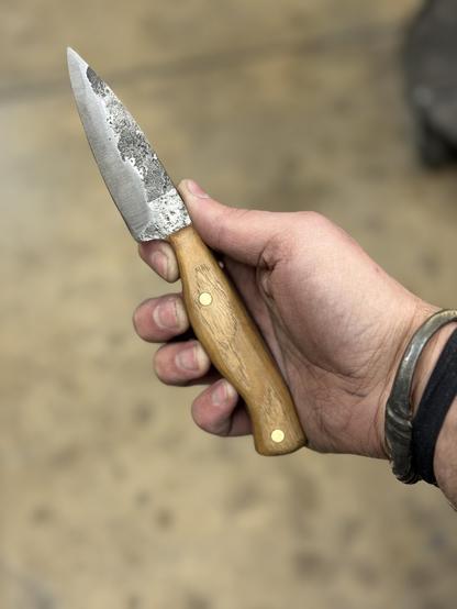 A small full tang hand forged knife with an ash wood handle and brass pins. It’s being held in front of the camera by my extremely dirty from forging hand.
