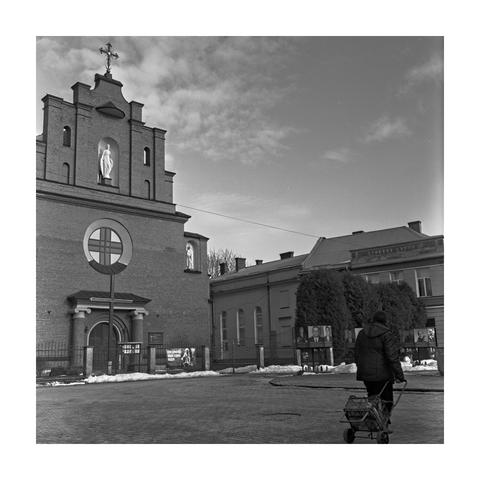 A sharp, square-format black and white photograph of a brick church in Horodok, Ukraine, captured on a Rolleiflex 3.5F with Kodak T-Max 400 film. The church features a prominent circular window with a cross and a statue of the Virgin Mary in an alcove. To the right, a local school building is visible. In the foreground, a person is seen from behind, pushing a small shopping cart across the paved square. The image displays exceptional detail and a rich tonal range from the bright, cloudy sky to the subtle textures of the brickwork and pavement.