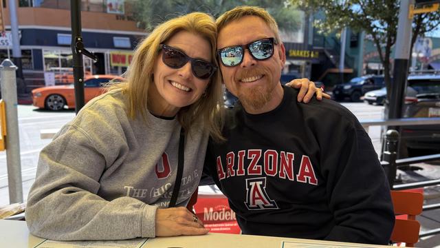 A smiling woman and man wearing sunglasses sitting closely together outdoors on a sunny day.
