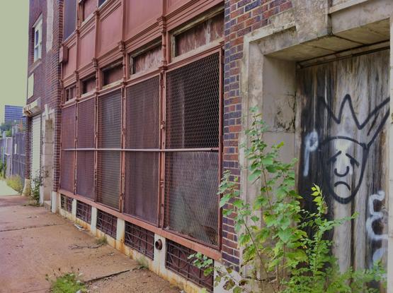 A brick building with metal protective grates over the windows and graffiti spray painted over plywood covering the door in Newark, NJ.