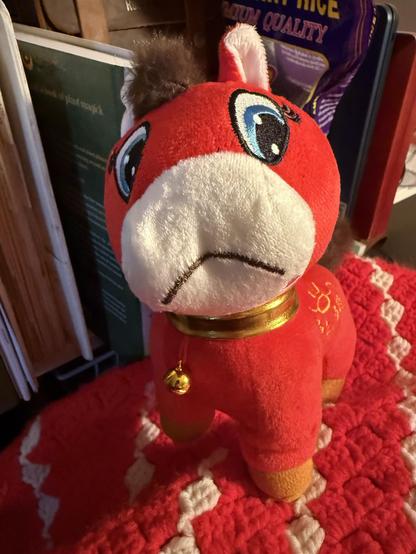 One of the iconic incorrectly-sewn red New Year's horse plushies with its muzzle on upside down so it looks like it's frowning. Me too, celebratory horse. Me too. It's sitting on a red and white crocheted afghan next to a bookshelf with a big packet of red rice berry rice on it.
