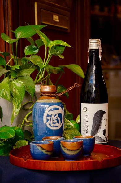 Composizione elegante e suggestiva che ritrae l’arte della tradizione giapponese: una bottiglia di sake Moonsoon, con un’etichetta minimalista che raffigura un volto femminile, affiancata da un antico vaso in ceramica blu con ideogramma bianco.
Sul vassoio in legno rosso, tre piccole tazze in ceramica smaltata, dai toni blu e terracotta, attendono di essere riempite. Sullo sfondo, una pianta di pothos con foglie rigogliose e variegate aggiunge un tocco di freschezza naturale, creando un’atmosfera di armonia tra natura, arte e ritualità.