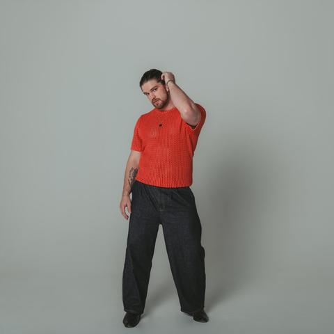 A white man brushes his brown hair back over his ear. He wears a bright red sweater with the sleeves rolled up, boxy blue jeans, and stands against a gray background. He has a light stubble. His head is titled and he stares directly into the camera.