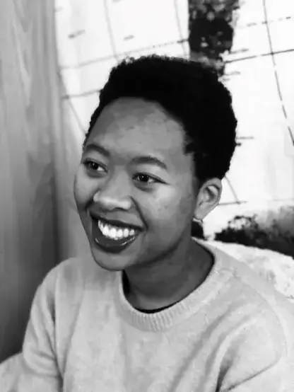 Black-and-white photo of Kemi Ashing-Giwa, a Black woman with a natural worn short on the sides and tall on the top. She wears a fine-gauge crew-neck sweater in a light tone and lip gloss. They sit on a chair or sofa with an upholstered back, in front of a light source (a curved window with panes, or perhaps a translucent tent?). She is turned with her face in three-quarters view as she looks attentively toward something or someone off-camera. Their eyes are focused, creased at the corners as part of their warm, broad smile.