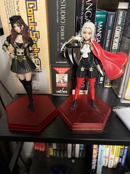 Figurines of Dorothea and Edelgard from Fire Emblem Three Houses, both on a bookshelf. Both are in their pre-timeskip outfits.