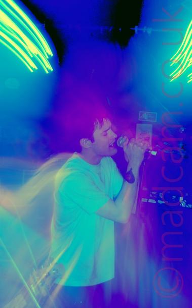 Long exposure of a man with dark hair, wearing a white t-shirt and a blue pair of jeans, holding a microphone in both hands as he sings.
A long exposure shot, starting zoomed out then zooming in, with no flash.
The result has clouds of blue and purple lights around his head, with streaks of green lights in both top corners, with secondary images of the singer behind him.
