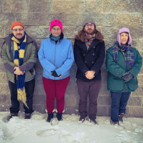 Four people in winter gear stand against a gray stone wall. There is snow on the ground. They look directly into the camera.