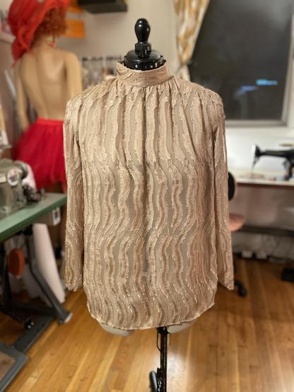 Front view of a bronze silk chiffon blouse, unbroken at the front and gathered around the shoulders and neck, with a high banded collar. The silk is "figured" with woven on squiggles of frayed silk satin and jacquard. It is on a dressform with a black stand and neck.