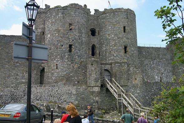 A large stone fortress with two round towers dominates the scene, its weathered grey walls rising above a wooden staircase leading to an arched doorway. Small, narrow windows punctuate the thick walls, and a flag flies faintly above the battlements. A lamppost with blank directional signs stands in the foreground beside parked cars, while several people walk and gather near the entrance, some appearing to be visitors or tourists. The surrounding stone wall extends to either side, and a small tree with green leaves frames part of the right edge against a blue sky with scattered clouds.