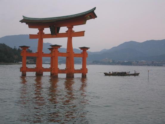 Heute regnet es. Direkt vor der Küste in Hiroshima Miyajima steht dieses Japanische Tor im flachen Wasser. Seine rot lackierten Säulen kontrastieren mit der grauen Bergkette im Hintergrund. Das Tor hat ein breites, dunkles Dach das an beiden Enden etwas nach oben schwingt. Zwischen dem Dach und dem Sturz des Tores prangt das Familienwappen der Familie, die das Kloster Miyajima baute. Rechts neben dem Tor schickt sich ein flaches, langes Boot mit vielen Menschen an, durch das Tor zu fahren.