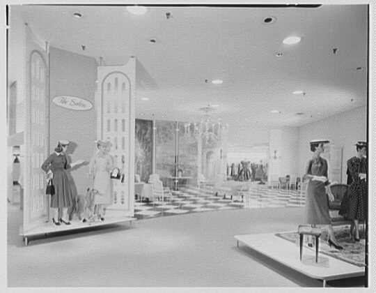 The image depicts a vintage interior of a department store, specifically focusing on the fashion section. There are large display windows filled with mannequins wearing dresses and hats in an organized manner to showcase clothing styles from that era.

In one window, there's a woman dressed elegantly standing next to another female figure which appears smaller due to perspective or scale representation. She is likely assisting her by holding the dress for better presentation of how it fits on the mannequin. In front of this display, an oval-shaped sign reads "The Salon."

On the opposite side, there are two more figures in a similar fashion setup with another woman standing beside one and possibly providing styling advice or assistance.

In the background, we see rows of other clothing items displayed neatly across shelves on various heights within the store. The flooring is checkered, indicating an older design typical for such establishments at that time period. Overhead fluorescent lighting illuminates the space evenly. There are a few customers visible in the distance and what seems to be staff wearing hats indicative of professional attire.

This image captures a moment from "Salon 1956" as mentioned in its source attribution, likely representing fashion trends or styles popular during that time at Lord & Taylor's department store located on Long Island. The company  [...]