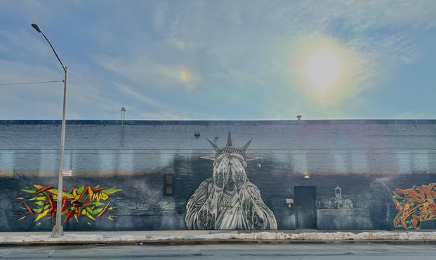 A wall of an industrial building in the south Bronx shows a wall mural of the Statue of Liberty crying with her face in her palms. To the left and right are colorful graffiti. The sky is blue and partly sunny.