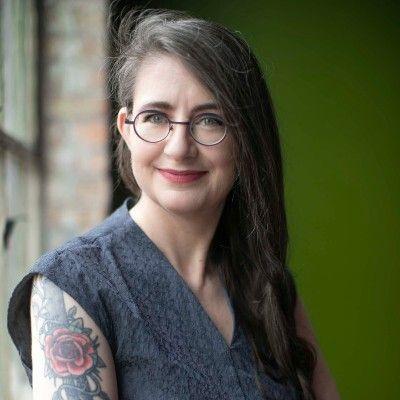 a picture of Heather Flanagan: a woman with round glasses, long dark hair with a direct gaze and a nice smile. She wears a dark sleeveless top showing a floral arm tattoo.