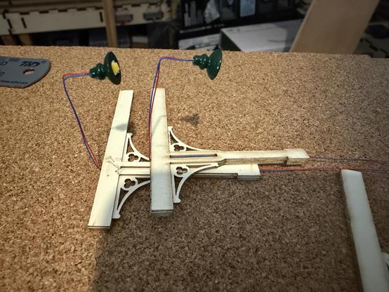 Two canopy supports lay on a sheet of cork. Both supports have the wires of a lamp running from the bottom through the middle, the wire bends to the right at the top, ready to be fixed to the crossmember when that is added. 

The wire will go through a small hole in the platform and from there to a 12V supply via a resistor.