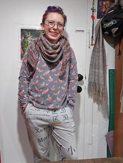Quinn standing by the door in the clothes described in the post, with comet earrings and cosmic pendant, in the background are a painting of Portland Frog, bike helmet, cutting boards for sewing, and woven scarf
