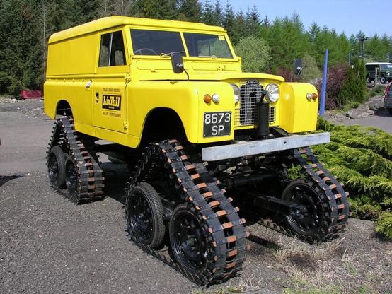 A beautifully preserved tracked Land Rover. The image shows an early model yellow Land Rover. In place of wheels, it has sets of triangular tracks. The scene is in sunshine.