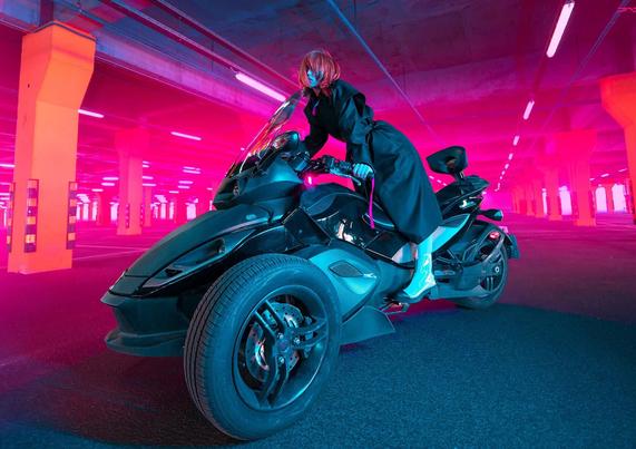 A person in a black coat poses on a sleek black three-wheeled motorcycle, set against a brightly lit, pink-themed parking garage.