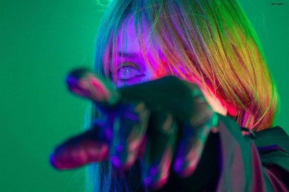 A close-up of a person with colorful lighting, wearing a black glove, reaching out with an expressive gaze and vibrant hair.