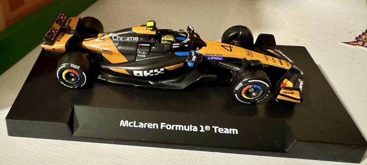 The image features a detailed model of a McLaren Formula 1 car, presented on a black base with the "McLaren Formula 1® Team" label. The car showcases a vibrant orange and black design with sponsor logos and the Number 4. Lando Norris’s helmet design - yellow and black in a kind of leopard print pattern - can be seen in the cockpit.