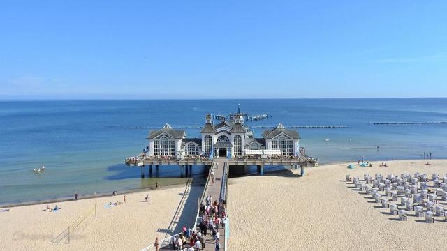 The image captures a vibrant seaside scene featuring a pier that extends into the ocean. The pier is an elegant structure with a large, ornate building at its end, characterized by multiple arches, large windows, and a classic architectural style. The building appears to be a restaurant or a pavilion, offering a scenic view of the sea.
The pier is connected to a sandy beach, where numerous people are seen walking, sunbathing, and enjoying the beach. The beach is lined with rows of beach chairs, likely for visitors to relax and enjoy the sun.
The ocean is calm, with gentle waves lapping against the shore. The sky is clear and blue, suggesting a beautiful, sunny day. The overall scene conveys a lively and inviting atmosphere, typical of a popular seaside destination.