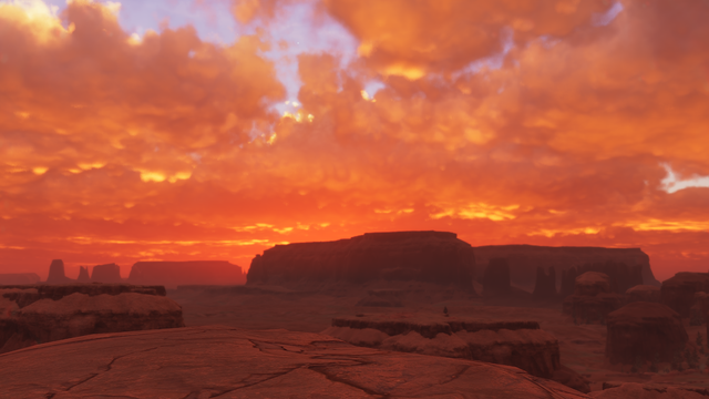 Elevated shot overlooking a clouded badlands desert landscape with numerous rocky buttes and mesas. The clouds obscure a low sun that is diffusing warm oranges, yellows, reds, and golds across the sky, while silhouetting the most distant rocky prominence
