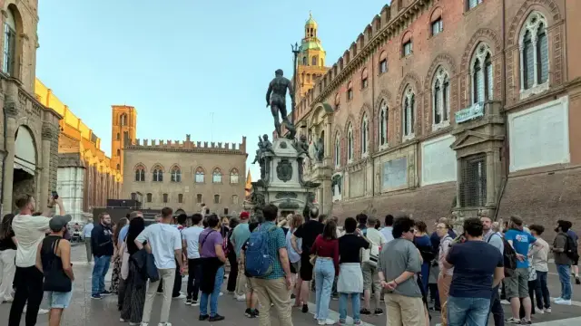 A photo taken during the guided city tour in Bologna. A group of people from PyCon Italia stands in Piazza del Nettuno, looking up and listening to the guide.