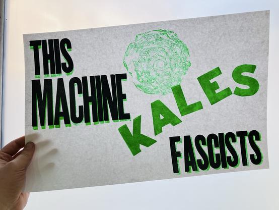 Photo of a letterpress print on white paper with black and neon green ink. It says "this machine kales fascists", with "kales" in green and the other words in black with green offset shadow behind, print held up in front of a frosted glass door with some light coming through. There is a large round print made by inking a cross-section of actual red cabbage, showing its layered packed leaves' texture.