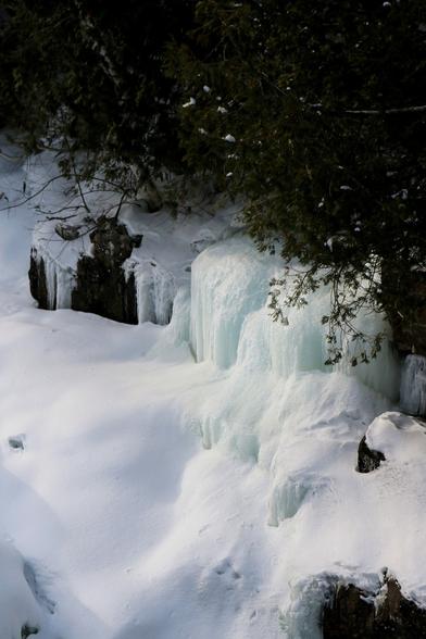 Photograph of ice formed by a small stream flowing over rocks on the bank of a river, freezing on contact with the air and forming large white ice cubes with a slightly turquoise tint, contrasting with the very white bluish snow covering the riverbed, under the green foliage of cedars.

Photographie de la glace formée par un petit ruisseau s'écoulant sur des rochers sur la rive d'une rivière, gelant au contact de l'air et formant de grands glaçons blancs un peu turquoises, contrastant avec la neige très blanche bleutées recouvrant le lit de la rivière, sous le feuillage vert de cèdres.