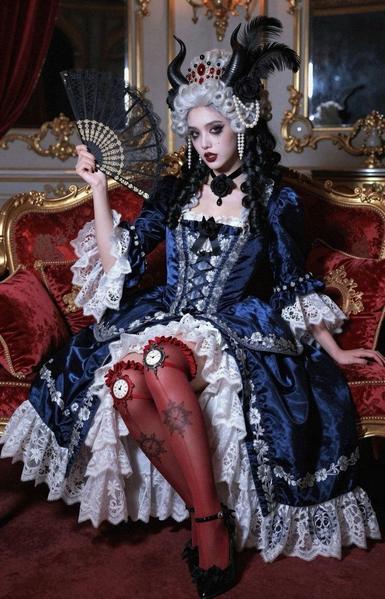 A woman in an elaborate blue and white dress, styled like 18th-century fashion, sits on a red velvet chair. She holds a black lace fan and wears dramatic makeup, including dark lipstick and striking eye makeup. Her outfit features red stockings with clock patterns