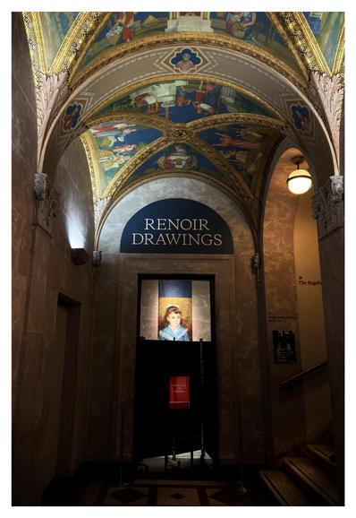 A hallway inside the Morgan Library and Museum in New York. Marble walls rise up to a groin-vaulted ceiling with elaborate ornamentation and Renaissance-themed imagery. An overhead lamp illuminates a stairway leading off to the right. In the middle of the photo is a doorway under a lunette with large text that reads “RENOIR DRAWINGS.” Through the doorway we can see a banner with an artwork reproduction hanging on a far wall: Pierre-Auguste Renoir’s “Portrait of a Girl (Elisabeth Maître),” featuring a brown-haired girl in a blue dress. The lower part of the banner is blocked from view by dark folding screens and a small red sign that says “INSTALLATION IN PROGRESS.” A rope barrier between two stanchions further discourages entry.