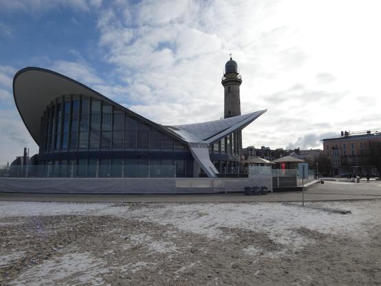 Der Teepott in Warnemünde, im Hintergrund der Leuchtturm und einige Gebäude. Vorne der Strand mit Schnee. Helle Wolken durch die die Sonne scheint.