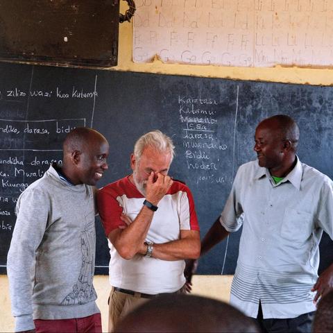 Tres hombres están conversando frente a una pizarra escrita con palabras en un aula.

En la imagen se observan tres hombres de pie, aparentemente en un entorno educativo, probablemente una escuela. La pared detrás de ellos tiene una pizarra negra con varios textos escritos a mano en un idioma que parece ser swahili o similar, con palabras organizadas en listas o columnas. Encima de la pizarra hay un cartel con letras del alfabeto escritas a mano. Los hombres visten ropa casual: uno lleva una camiseta blanca con mangas rojas, otro un suéter gris y el tercero una camisa clara. La atmósfera es amable, con la persona del centro llorando y tapándose la cara con la mano de forma emocionada, mientras las otras dos personas le miran con una sonrisa.