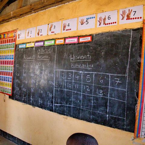 La imagen muestra un aula con una pizarra llena de lecciones escritas en suajili, enfocadas en números, días de la semana y fonética.

En detalle, se observa una pizarra negra donde están escritas palabras y tablas relacionadas con el conteo ("HISABATI" que significa matemáticas) y fonética de vocales y sílabas en suajili. Arriba de la pizarra hay tarjetas con imágenes de manos mostrando números del 0 al 7, junto con puntos rojos que refuerzan la enseñanza visual del conteo. También hay un cartel colorido con números del 1 al 100 pegado en la pared a la izquierda. Encima de la pizarra están indicados los días de la semana en suajili (Jumapili, Jumamosi, Ijumaa, etc.). La atmósfera es educativa y organizada, pensada para facilitar el aprendizaje básico de matemáticas y lenguaje.