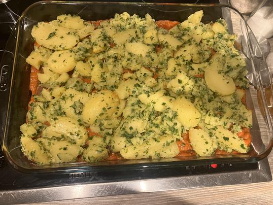 A glass baking dish containing a layered dish with a base of veggie sausage, baked beans and a topping of mashed potatoes mixed with herbs, showing chunks of potato on top.