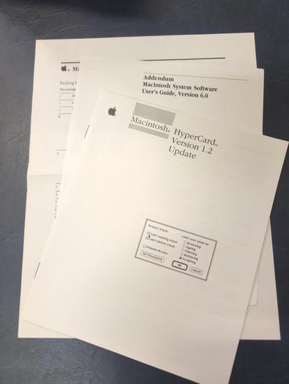 A stapled booklet on top of two other sheets of paper on a blue table. Printed in black on the cover of the booklet is the Apple logo and Macintosh HyperCard Version 1.2 Update. Below that is a picture of a Mac window with checkboxes and radio buttons.