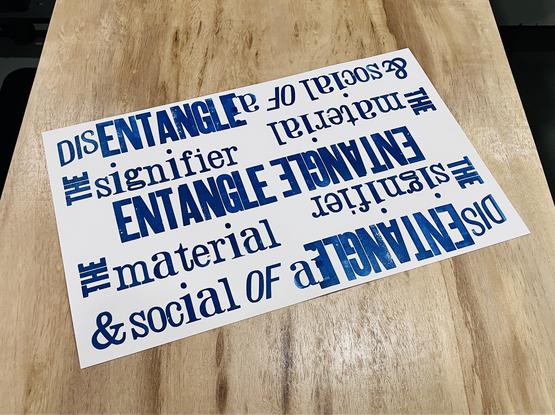 Photo of a letterpress print on white paper with blue inked text saying "Entangle the signifier, entangle the material & social of ai"; the print has been turned 180 degrees to print the same text from the other direction upside down