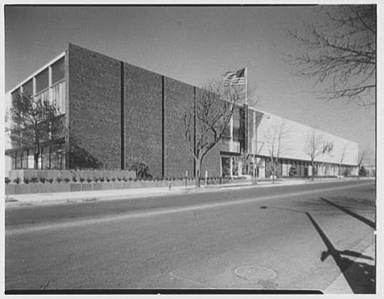 The image shows a black and white photograph of an old brick building with large windows, which appears to be from the 1950s. The American flag is displayed prominently on top right corner of the facade, indicating it might have been taken in the United States during that era.

There are leafless trees visible along the side walk, suggesting either a winter or early spring season when most trees have not yet blossomed. A street runs parallel to one end of the building with a manhole cover on its surface and no cars parked alongside suggest it may be less busy at this time or possibly from an earlier era.

A shadow falls across part of the sidewalk, likely caused by either early morning sun or late afternoon light casting over the area in question. The lack of modern elements like street lamps hints that the photograph could have been taken a few decades ago when such lighting was not as prevalent on city streets for safety reasons and aesthetics.

The overall impression is one of an urban environment with commercial buildings, possibly during midday judging by the length of shadows observed which suggests it's neither early morning nor late evening. The absence of people in this scene focuses attention solely upon architectural details and historical context provided by style and condition of construction materials used.