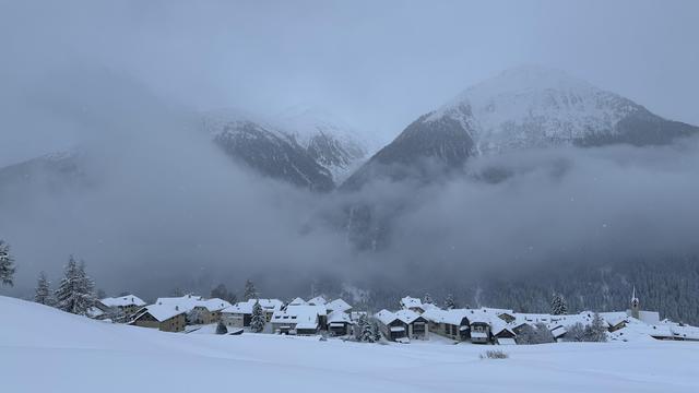 View of the snow-covered Engadine village of Guarda seen from above across snowy fields. The valley beyond the village is filled with low clouds which partially obscure the mountains rising on the opposite side.